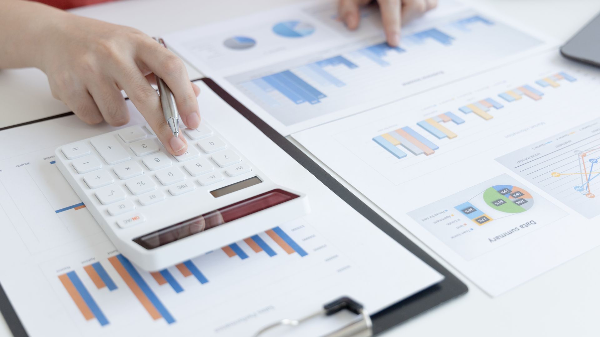Hands using a calculator over printed financial charts and bar graphs on a desk, with a smartphone nearby.