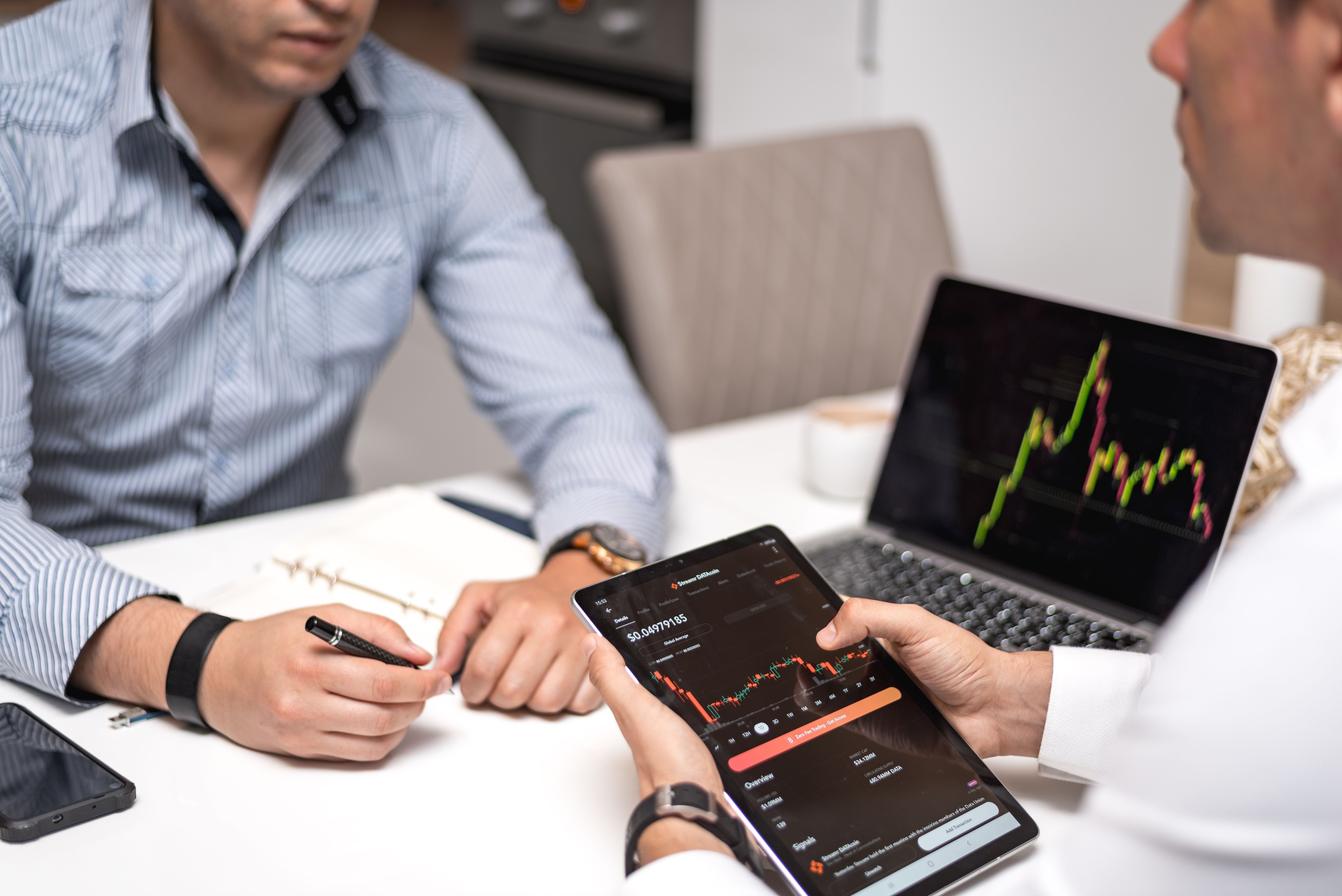 Two people at a meeting table, one holding a tablet displaying a cryptocurrency trading app with candlestick charts, the other holding a pen. A laptop showing a stock chart is visible in the background.