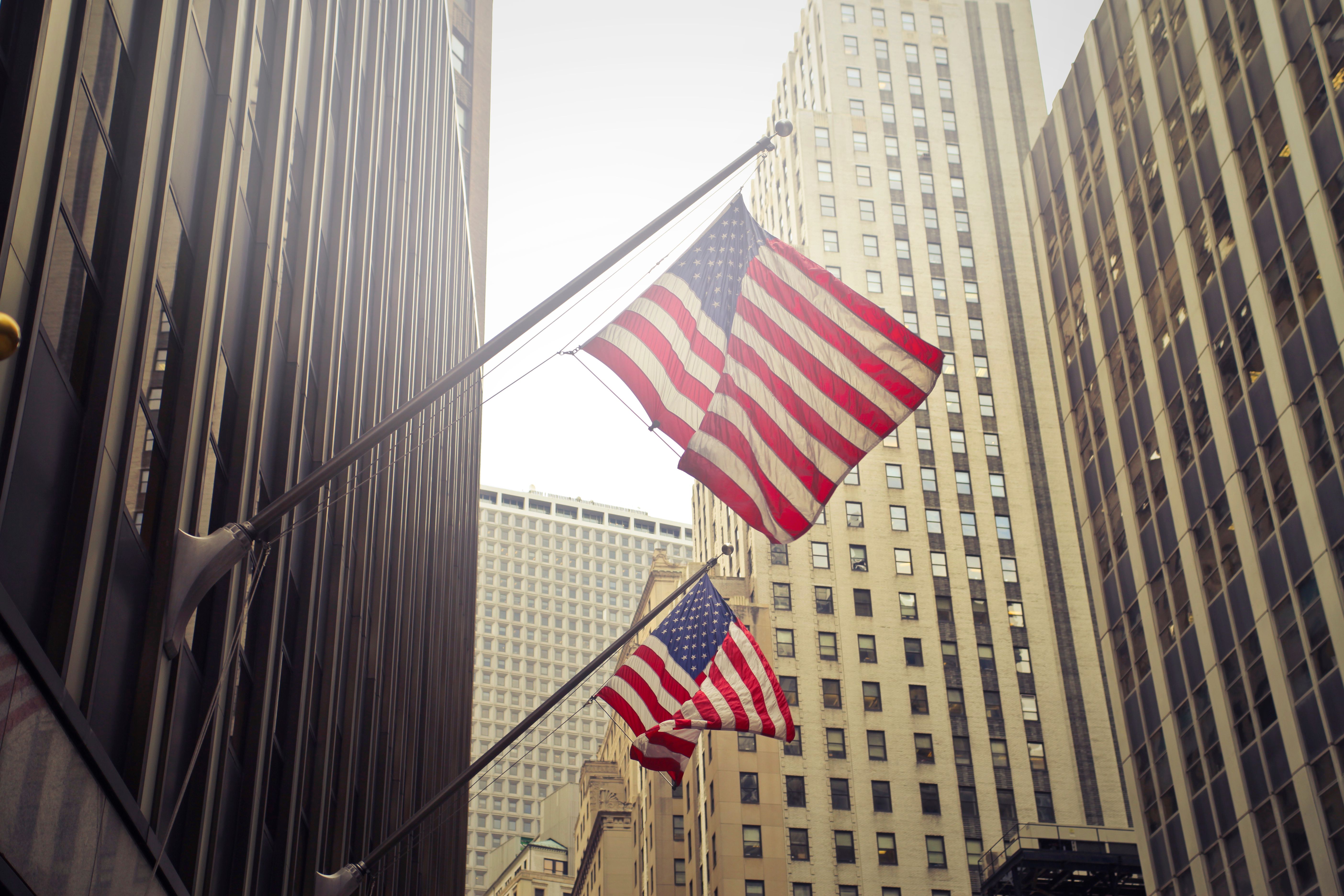 Two American flags on angled poles mounted to a building facade, seen from street level between tall office skyscrapers in a dense city financial district.