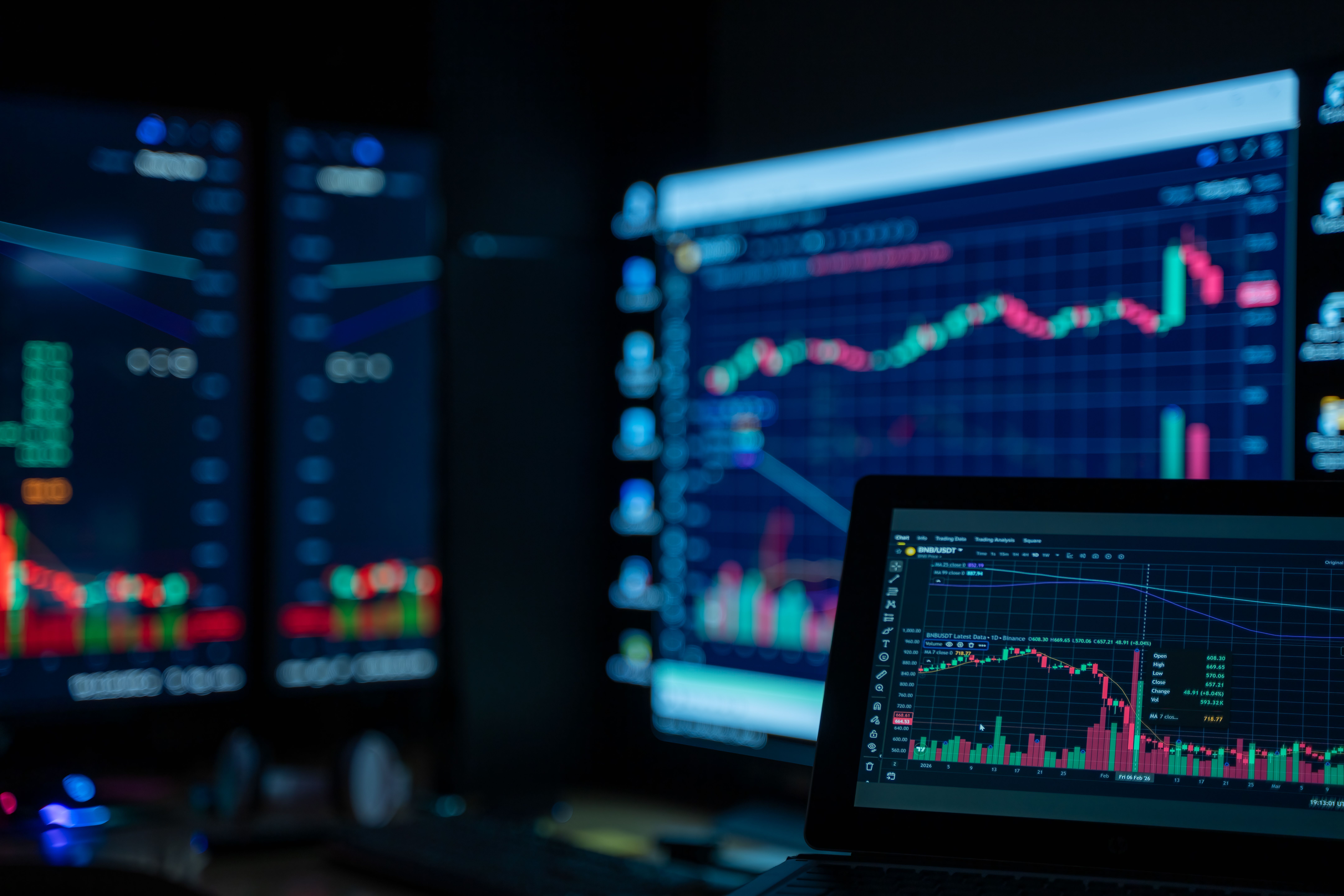 Laptop in a dark room showing a BNB/USDT cryptocurrency candlestick chart with volume bars, set against blurred monitors displaying more red and green trading charts.